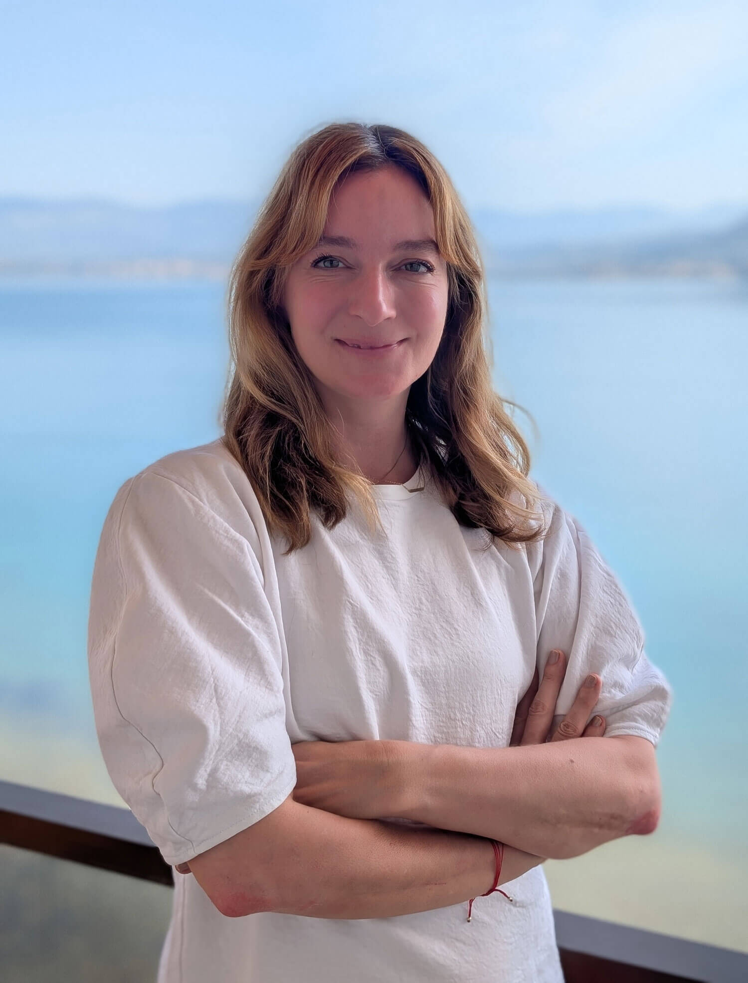 Foto von Kristina Roumeliotis, die vor dem Meer an einem Geländer steht und in die Kamera lächelt